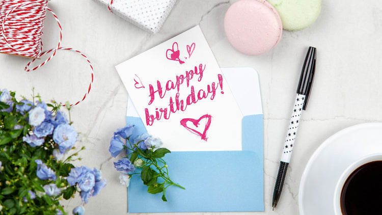 Birthday greeting card in a blue envelope with flowers, ribbon, macarons, a pen and coffee on a tabletop